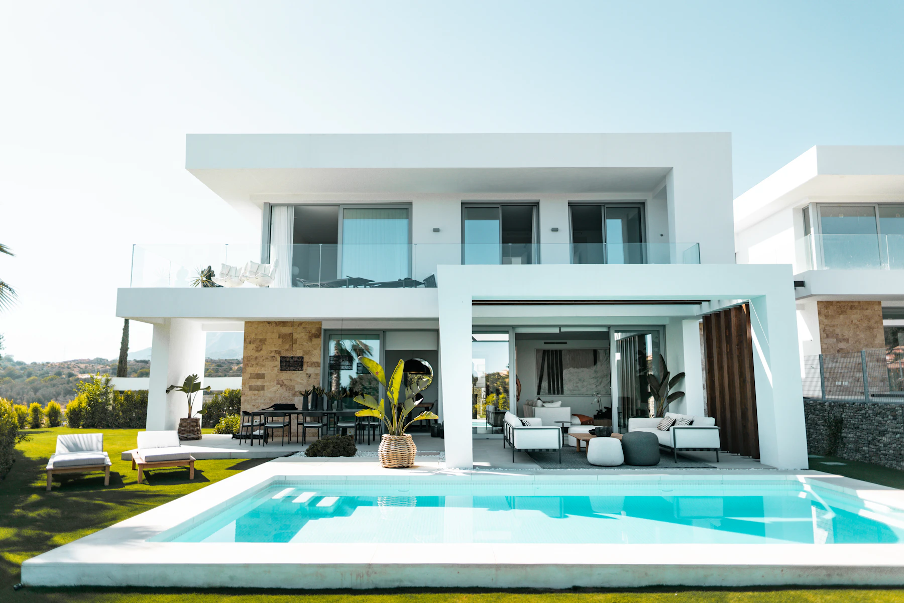 Completed luxury villa at dusk: white cantilevered upper storey over full-height
             glazing, stone-clad walls, an infinity pool and planted terrace